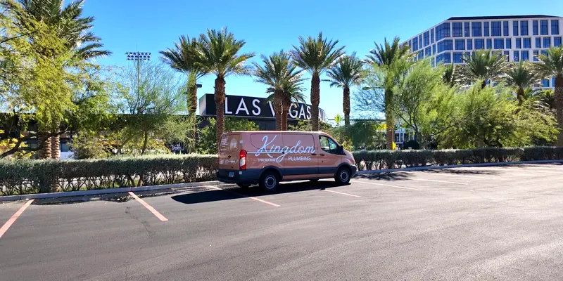 Tan Kingdom Plumbing van parked diagonally across red stall lines beside a hedge of palms with a partial LAS VEGAS sign and office tower behind