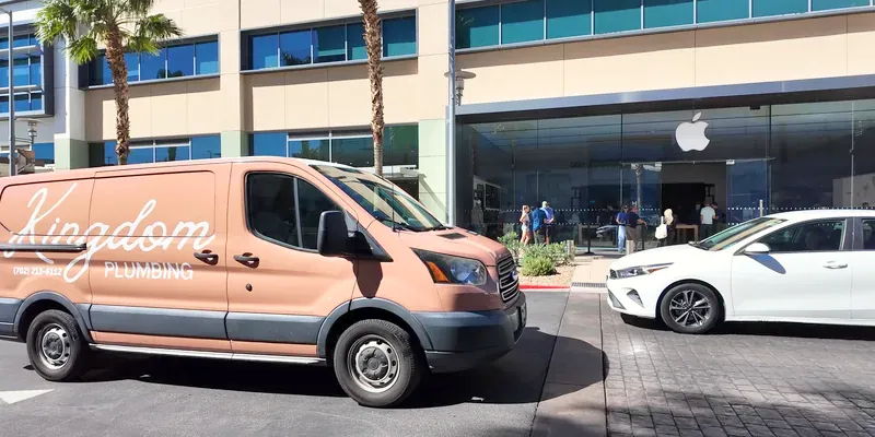 Kingdom Plumbing van nose-to-nose with a white car on the Downtown Summerlin pavers in front of the Apple Store and twin palm trees